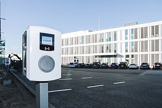 Eine weiße Ladestation für Elektrofahrzeuge steht auf einem Parkplatz. Im Hintergrund sind mehrere geparkte Autos und ein modernes Gebäude mit großen Fenstern zu sehen, unter klarem Himmel.