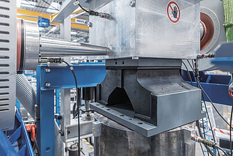 Foto einer großen metallischen Prüfmaschine mit blauen und silbernen Komponenten. In der Mitte befindet sich ein massives Bauteil mit einer großen Öffnung, das auf einer Plattform eingespannt ist.
