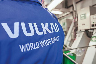 Rückseite einer blauen Jacke mit weißem Aufdruck ‚VULKAN WORLD WIDE SERVICE‘, im Hintergrund unscharfe Maschinen.
