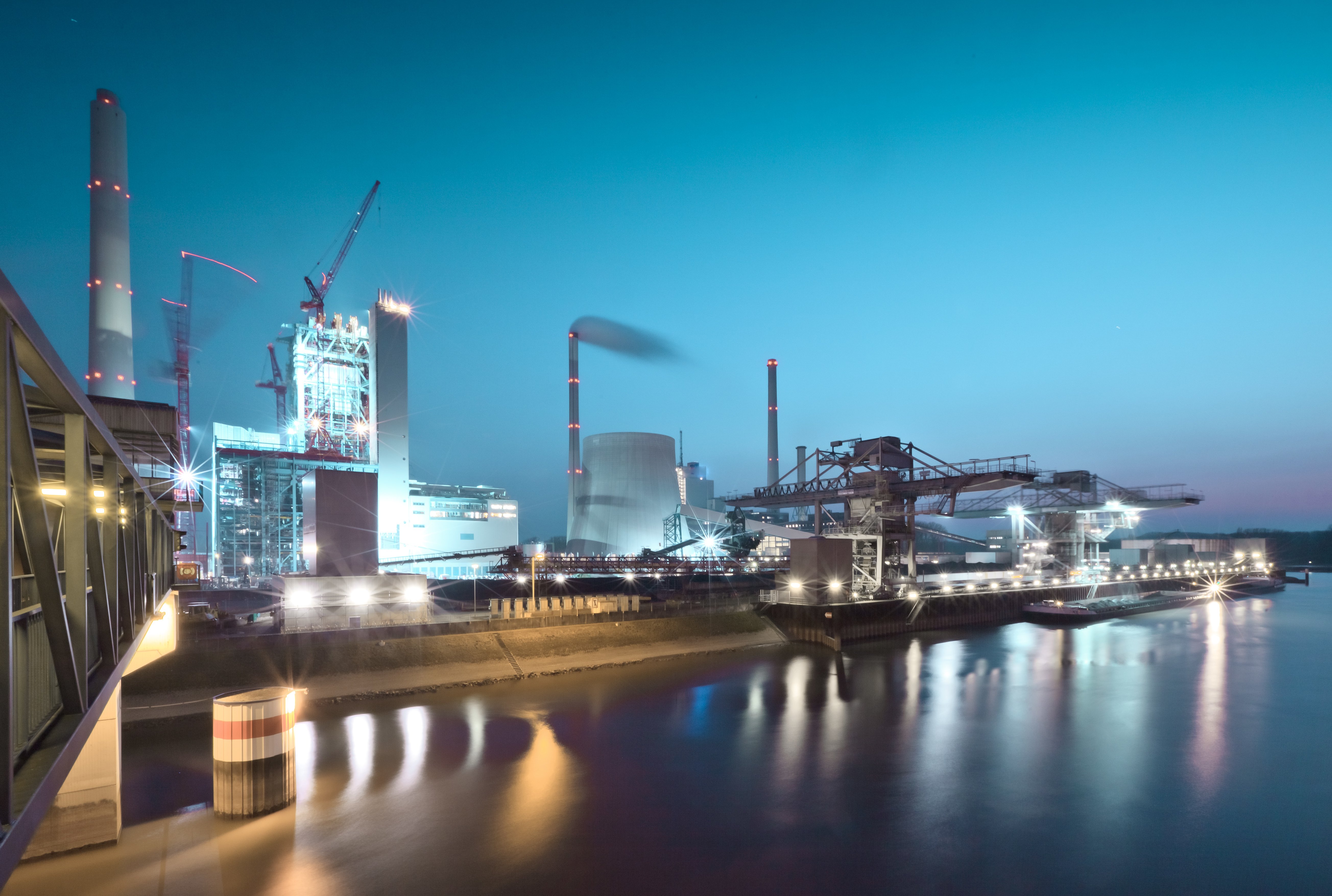 Industriegebiet bei Dämmerung mit beleuchteten Gebäuden, Kränen und Schornsteinen. Spiegelungen im angrenzenden Gewässer sichtbar.