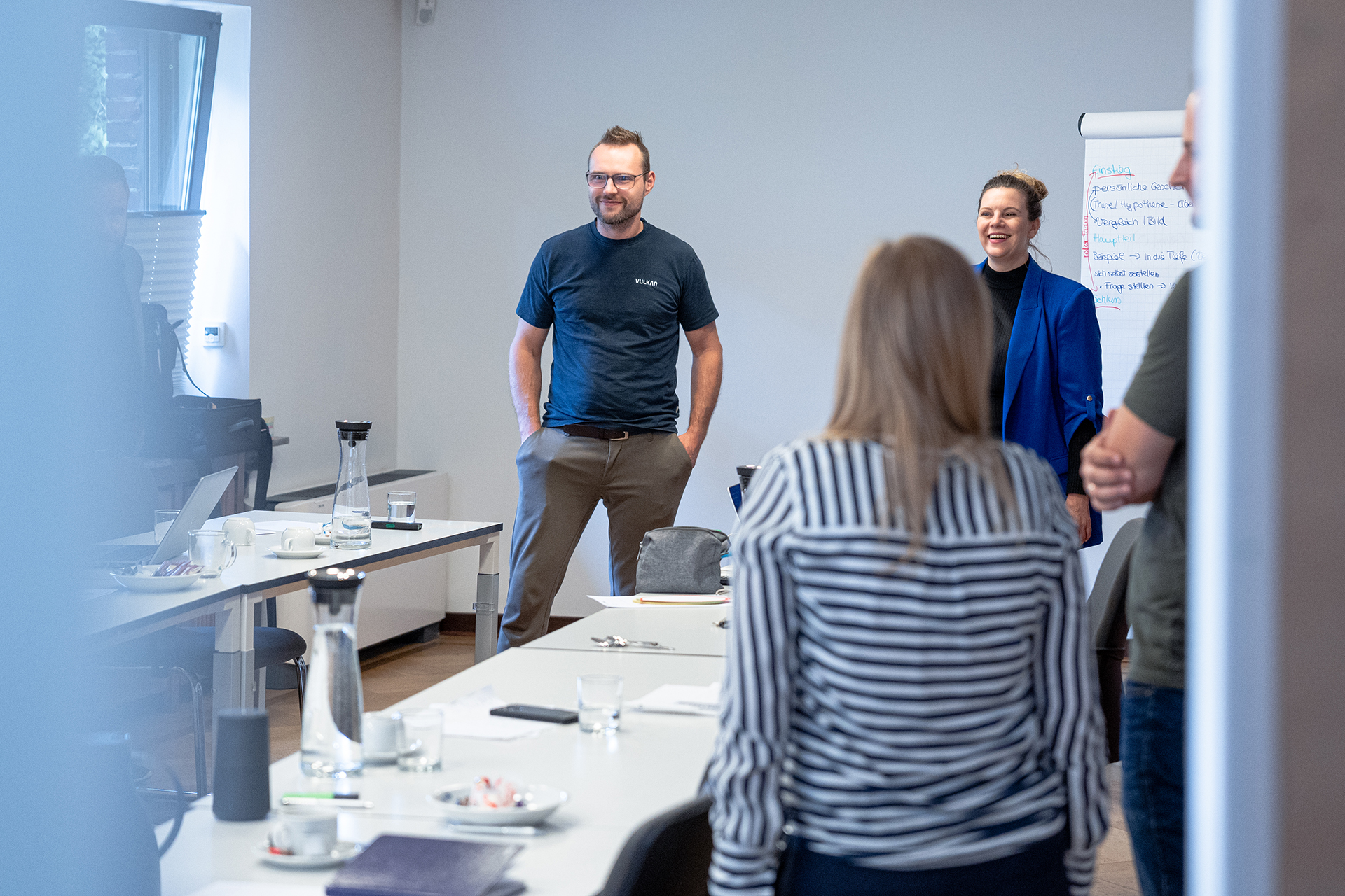 Eine Gruppe von vier Personen (3 Männer, 1 Frau) steht in einem Besprechungsraum. Eine Person ist mit dem Rücken zur Kamera zu sehen, drei Personen blicken in Richtung Kamera. Auf dem Tisch liegen verschiedene Gegenstände, darunter Papier, ein Laptop und ein Kaffeebecher. Im Hintergrund befindet sich ein Whiteboard oder Flipchart.