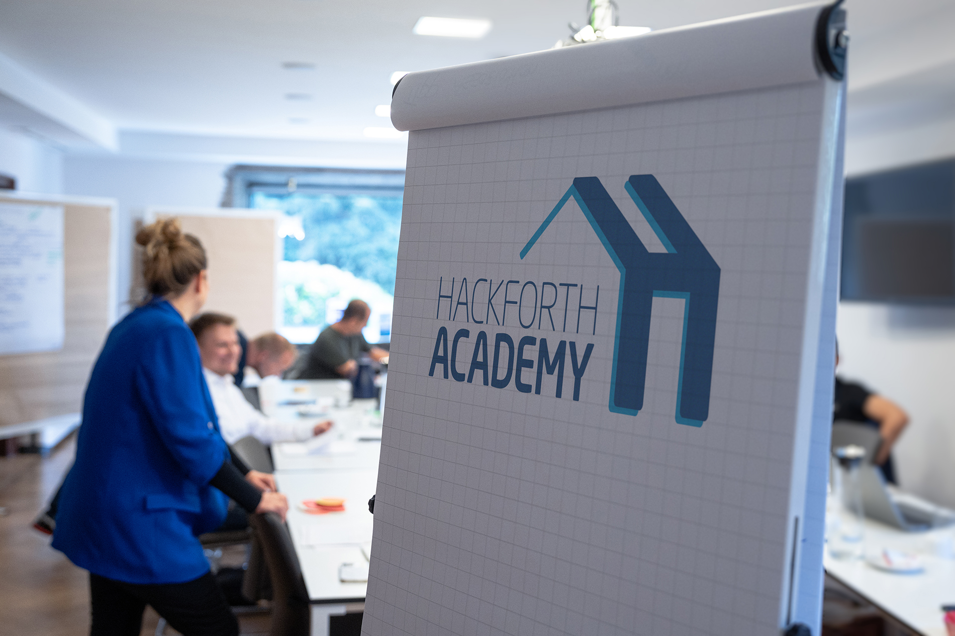 Eine Gruppe von Personen befindet sich in einem Besprechungsraum. Im Vordergrund steht ein Flipchart auf dem das Hackforth Academy Logo zu sehen ist.