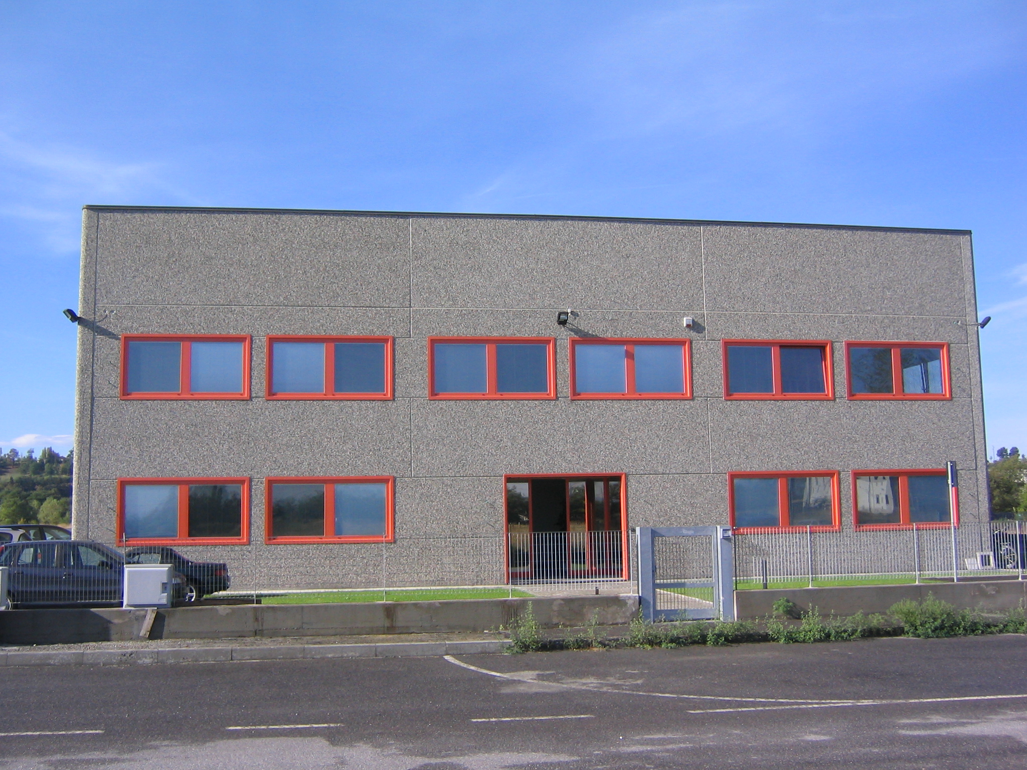 Ein zweigeschossiges Gebäude mit einer Fassade aus grauen Steinen und roten Fensterrahmen. Vor dem Gebäude befinden sich ein gepflasterter Bereich, einige Gegenstände und ein paar Pflanzen. Der Himmel ist blau.