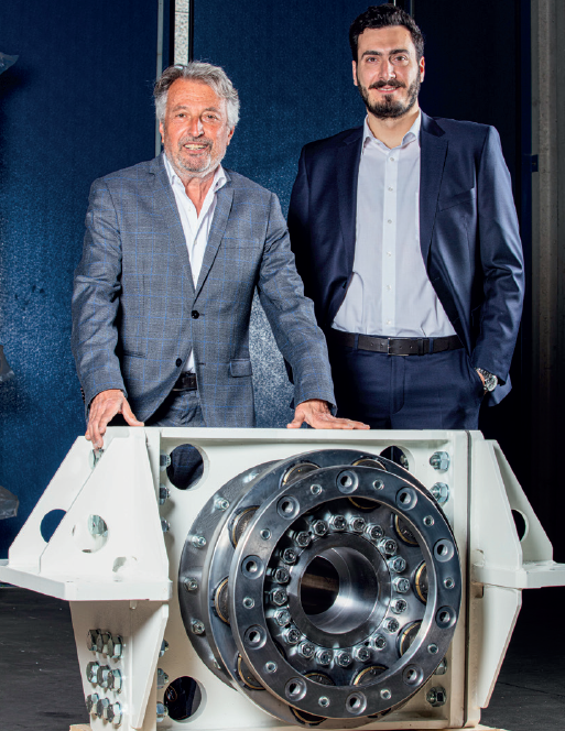Zwei Personen in Anzügen stehen hinter einem großen technischen Bauteil mit einer runden Metallkomponente in der Mitte. Der Hintergrund ist dunkelblau, und der Boden ist grau.