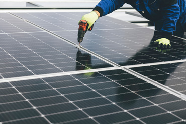 Eine Person arbeitet mit einem Akkuschrauber an der Oberfläche von Solarpanels. Die Person trägt gelbe Handschuhe und blaue Kleidung.